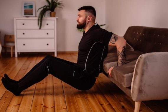 A bearded man in black fitness attire exercising at home, showcasing arm tattoos and a healthy lifestyle.